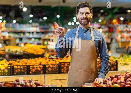 Employé de l'épicerie dans le tablier debout dans la section des fruits et légumes pointant le doigt. Fruits et légumes lumineux sur l'affichage créant un fond coloré. Image du service à la clientèle, de la vente au détail, de l'alimentation et du marché. Banque D'Images