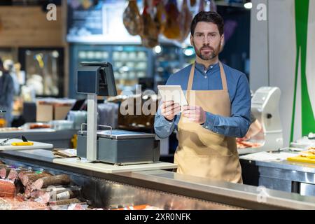 Boucher mâle dans le tablier en utilisant la tablette derrière le comptoir à la boutique de charcuterie. Affichage de différentes viandes et saucisses visibles. Transformation numérique dans le commerce de détail traditionnel Banque D'Images