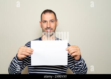 Un homme hispanique barbu dans les années 40 montre une feuille de papier blanche à la caméra sur un fond beige. Zone publicitaire. Banque D'Images