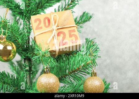 Boîte cadeau avec 2025 écrit en rouge sur le dessus de l'arbre de Noël décoré avec des boules dorées devant un fond gris. Gros plan. Banque D'Images