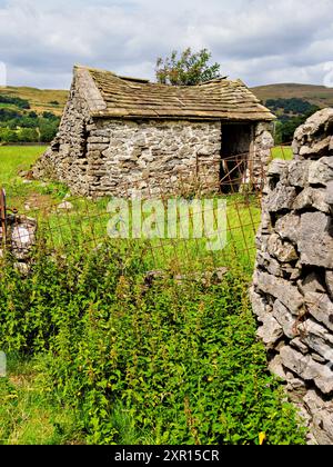 Une grange en pierre rustique se dresse dans un champ entouré de verdure luxuriante et de collines ondulantes sous un ciel nuageux. Banque D'Images