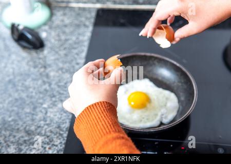 Vue aérienne des mains d'une femme plaçant un oeuf sur une poêle à frire sur une cuisinière à induction ouvrant ses coquilles Banque D'Images