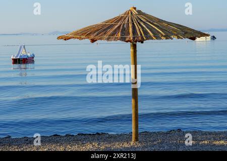 Parasol en rotin de plage de paille à la plage vide avec belle mer calme ou côte océanique sous le ciel bleu. Journée d'été relaxante. Voyage idyllique et vacances d'été Banque D'Images