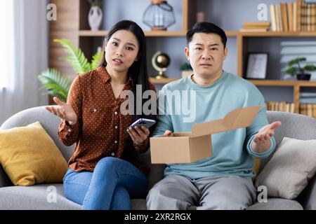 Couple asiatique assis sur le canapé se sentant frustré par la livraison du produit. Femme tenant smartphone et homme tenant boîte ouverte. Concept de questions d'achat en ligne, de service à la clientèle et de problèmes de livraison Banque D'Images