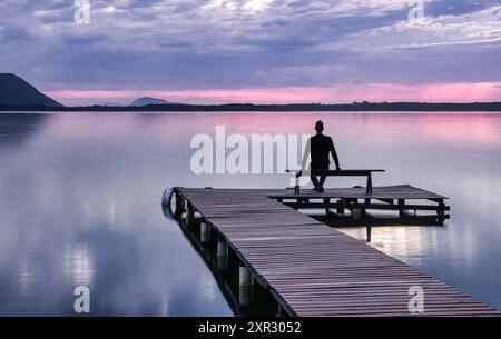 Homme assis sur un banc de quai en bois regardant le lever du soleil à Lagoa da Conceicao à Florianopolis - Brésil Banque D'Images