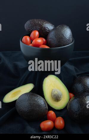 Tomates cerises rouges et avocats dans un bol noir avec un avocat coupé devant. Banque D'Images