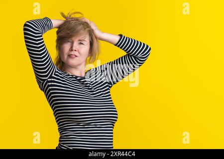 La femme mature aux cheveux blonds pose dans une chemise rayée à manches longues, levant ses mains derrière la tête et regardant la caméra. Le fond est jaune ensoleillé Banque D'Images