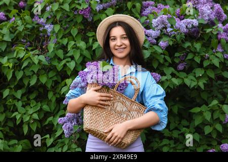 Belle jeune femme heureuse tenant un sac en osier avec bouquet de fleurs de lilas dans le parc le jour du printemps Banque D'Images