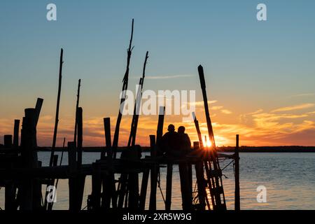 Couple d'hommes assis à regarder le coucher du soleil dans le port sur pilotis de Carrasqueira, à Comporta, Portugal. Banque D'Images