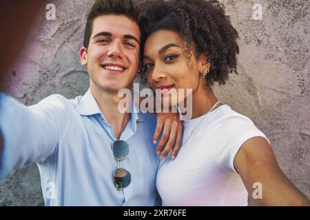 Jeune couple multiculturel partageant un moment romantique en prenant un selfie. Met en valeur la diversité, les relations modernes et la connexion joyeuse dans un urb Banque D'Images