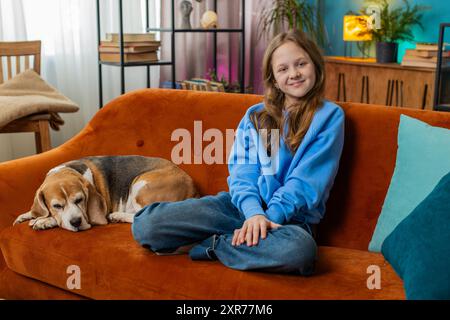 Portrait de jeune fille souriante avec chien beagle mignon assis sur le canapé orange. Heureux preteen femelle enfant amoureux des animaux passer du temps libre avec chien de compagnie dans le salon à la maison pendant le week-end. Banque D'Images