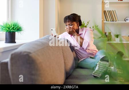 Surprise femme afro-américaine choquée à la maison obtenant des nouvelles au téléphone intelligent en état de choc Banque D'Images