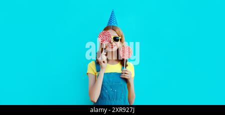 Vacances, célébration, événement. Joyeuse jeune femme rit célébrant s'amuser à une fête portant un chapeau d'anniversaire festif tenant sucette sur bleu Banque D'Images