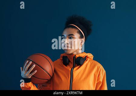 Jeune femme avec basket-ball et écouteurs sans fil posant en studio. Sportive confiante dans une tenue de fitness orange sur fond bleu regardant AWA Banque D'Images