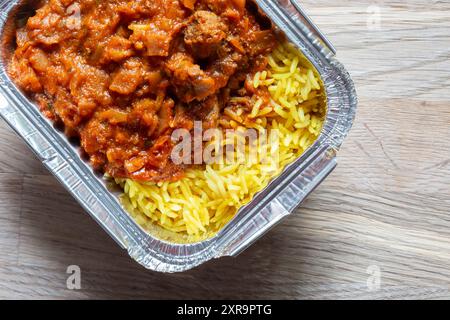 Agneau Rogan Josh curry indien avec riz pilau repas à emporter dans un emballage de plateau en aluminium. Sur fond de planche de bois Banque D'Images
