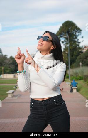 Belle jeune femme avec une bonne attitude souriante dans des vêtements décontractés se tient debout dans le parc pointant vers le ciel avec ses doigts, image verticale Banque D'Images