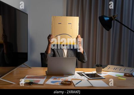 Portrait d'homme d'affaires couvrant son visage avec un masque de papier avec un visage triste dessiné dessus, mauvaise personne anonyme de dépression sullen négative. Banque D'Images