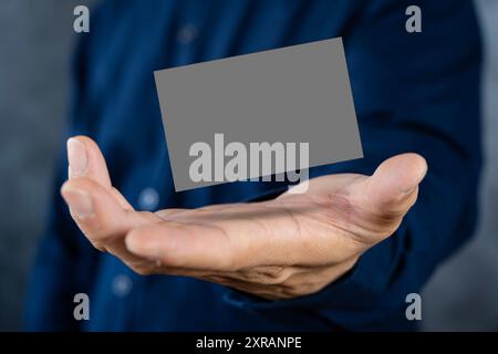 Homme tenant une carte de visite gris foncé contre un mur de béton main masculine mature tenant une carte de visite vierge devant la caméra. Backgroun flou Banque D'Images