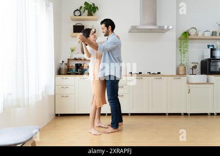 Couple heureux s'amusant dans la cuisine, dansant ensemble, célébrant la migration ou l'anniversaire, beau jeune mari tenant la main d'une belle épouse Banque D'Images