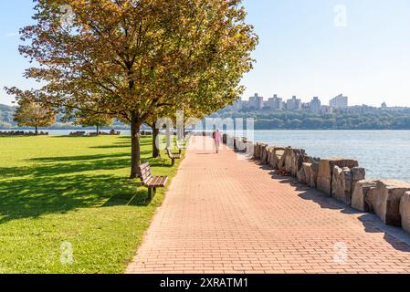 Femme faisant du jogging sur un chemin de briques bordé d'arbres et de bancs dans un parc au bord de la rivière par un matin d'automne clair. Banque D'Images