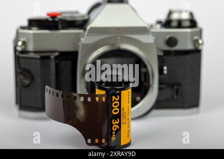 Vue détaillée d'un rouleau de film de 35 mm en mise au point, avec un vieil appareil photo sans lentille flou en arrière-plan. Le fond blanc souligne la sensation classique. Banque D'Images