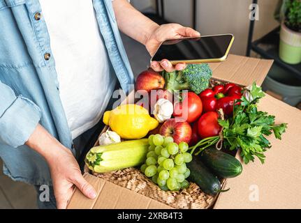 Commande en ligne de légumes et de fruits cultivés à la ferme. Téléphone portable avec écran blanc dans la main de la personne féminine et boîte en carton avec des légumes frais. Banque D'Images