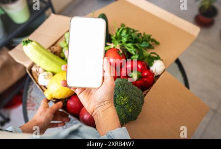 Commande en ligne de légumes et de fruits cultivés à la ferme. Téléphone portable avec écran blanc dans la main de la personne féminine et boîte en carton avec des légumes frais. Banque D'Images