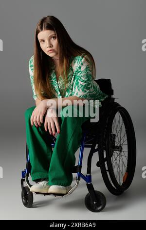 Une jeune femme portant des vêtements verts est assise dans un fauteuil roulant sur un fond gris. Banque D'Images