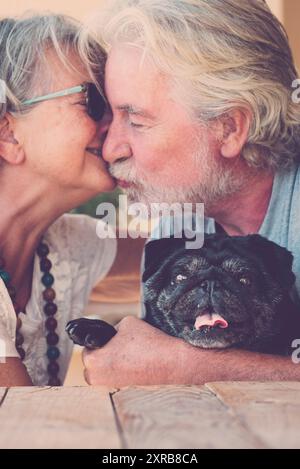 Les personnes âgées de race blanche couple baiser et l'amour avec le chien noir pug au milieu - portrait de l'homme heureux et la femme âgés style de vie à la maison - animal drôle et vieux propriétaire apprécient l'amitié et la relation Banque D'Images