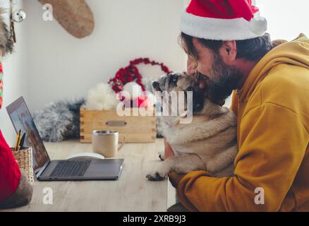 Fête de Noël à la maison avec un seul homme en vidéoconférence sur ordinateur portable qui embrasse son adorable chien. Concept de personnes appréciant décembre à la maison avec des décorations de Noël et meilleur ami Banque D'Images