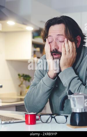 Homme fatigué à la maison le matin réveil activité touchant le visage et étirant après une mauvaise nuit de repos. Concept de travail en ligne de café et d'ordinateur. Une personne et l'état de santé de l'insomnie. Avant l'activité du travail Banque D'Images