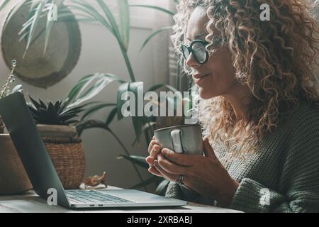 Une femme sereine avec des cheveux bouclés sur l'affichage d'ordinateur portable et boire du café ou du thé. Des gens sains modernes travaillant à la maison avec des plantes d'intérieur de jardins verts en arrière-plan. L'humeur verte colore le style de vie des gens Banque D'Images
