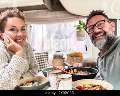 Heureux couple mangeant ensemble à l'intérieur d'un camping-car camping-car vivant la vie vanlife et voyage aventure vacances. Vie nomade. Homme et femme joyeux prenant selfie et souriant à la caméra Banque D'Images