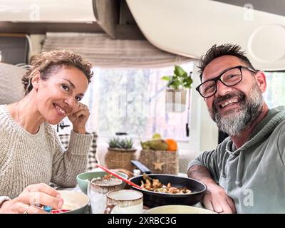 Heureux couple mangeant ensemble à l'intérieur d'un camping-car camping-car vivant la vie vanlife et voyage aventure vacances. Vie nomade. Homme et femme joyeux prenant selfie et souriant à la caméra Banque D'Images