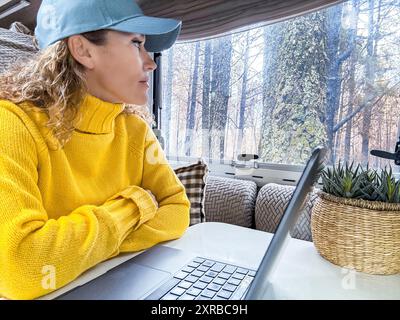 Belle femme en chapeau bleu et pull jaune regarde la fenêtre du camping-car devant le top pour ordinateur portable. Lady aime la forêt assise de son camping-car Banque D'Images