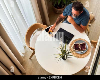 Vue supérieure au deuxième étage de l'homme ayant le temps de petit déjeuner à la maison seul en utilisant un ordinateur portable à la table. Chambre d'hôtel ou concept d'appartement confortable style de vie des gens modernes travaillant tôt vérifier les e-mails ou les notifications Banque D'Images
