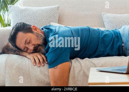 Un homme s'endort à la maison pour la sieste de l'après-midi couché sur le canapé avec ordinateur portable ouvert sur la table. Les personnes fatiguées dorment pendant la journée. Surmenage concept de stress santé de style de vie. Dormir sur le canapé Banque D'Images