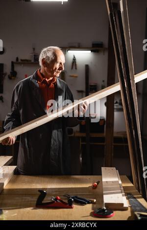 Un charpentier âgé debout dans son atelier de menuiserie, tenant une planche de bois dans ses mains. L'artisan chevronné est entouré de divers outils Banque D'Images