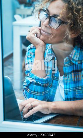Femme souriante en chemise à carreaux travaillant sur ordinateur portable tout en étant assis au bureau près de la fenêtre dans un laboratoire ou à la maison. Vision à travers la fenêtre. Dame adulte rêvant et pensant regardant à l'extérieur de la veuve Banque D'Images