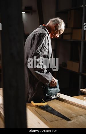 Menuisier âgé utilisant une scie sauteuse électrique pour couper des planches de bois découpées dans son atelier de menuiserie Banque D'Images