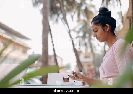 Petit déjeuner, déjeuner à l'air frais. Jeune femme indienne aime le dessert sucré, s'assoit à table dehors. Banque D'Images