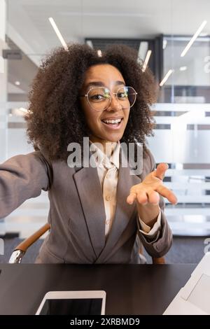Jeune femme d'affaires afro-américaine sur le lieu de travail à l'intérieur du bureau appel vidéo regardant la caméra pov. Professionnel en costume gris avec lunettes communiquant. Environnement de bureau moderne avec cloisons en verre Banque D'Images