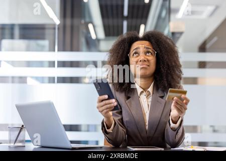 Jeune femme d'affaires afro-américaine se sentant frustrée au lieu de travail tenant le téléphone et la carte de crédit. Ordinateur portable sur le bureau dans un bureau moderne transmet le stress, les problèmes financiers et les transactions numériques Banque D'Images