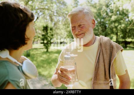 Homme aux cheveux gris tenant une bouteille d'eau et souriant à sa femme brune après avoir couru dans le parc Banque D'Images