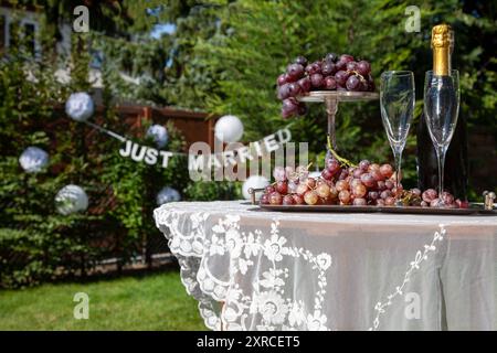 Au premier plan se trouve un arrangement avec 2 verres à champagne et une bouteille de champagne à côté de raisins rouges sur un plateau en argent, table avec nappe blanche dans un jardin, préparation pour un mariage dans le jardin, lettrage JUSTE marié dans le fond hors focus Banque D'Images