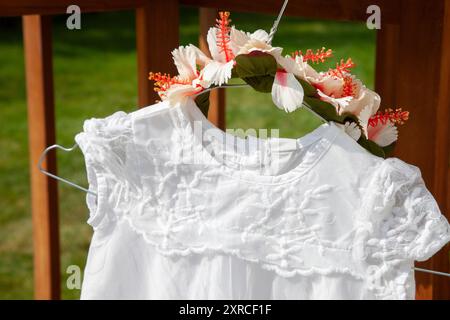 Gros plan d'une robe d'enfant blanche avec de la dentelle et une couronne de fleurs comme coiffe suspendue à un cintre en fil de fer sur une table, préparations pour un mariage, préparation de mariage à l'extérieur Banque D'Images