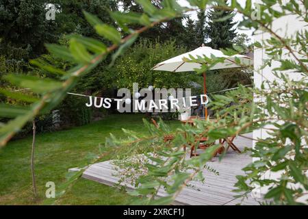 Premier plan flou avec des feuilles vertes d'un saule, focus sur une terrasse avec salon de jardin et lettres bannière JUSTE MARIÉ, préparation et décoration Banque D'Images