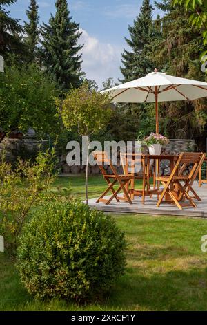 Salon de jardin en bois et parasol lumineux sur une terrasse en bois au dernier soleil de la journée, jardin verdoyant en fin d'été, soleil du soir Banque D'Images