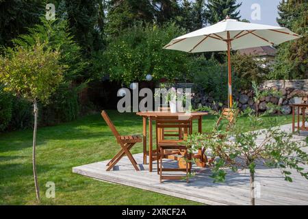 Au premier plan sont les feuilles vertes d'un jeune saule, en arrière-plan mobilier de jardin et parasol sur une terrasse en bois dans la dernière lumière du soleil de la journée, sièges dans le soleil du soir dans le jardin verdoyant à la fin de l'été, en arrière-plan brouillé le jardin vert Banque D'Images