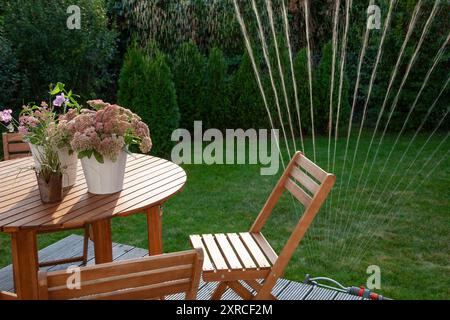 Fleurs en fleurs dans des pots blancs sur une table en bois, terrasse dans le jardin verdoyant, en arrière-plan un arroseur arrose la pelouse et les conifères verts, les jets d'eau brillent dans le contre-jour du soleil du soir et sont un rafraîchissement rafraîchissant Banque D'Images